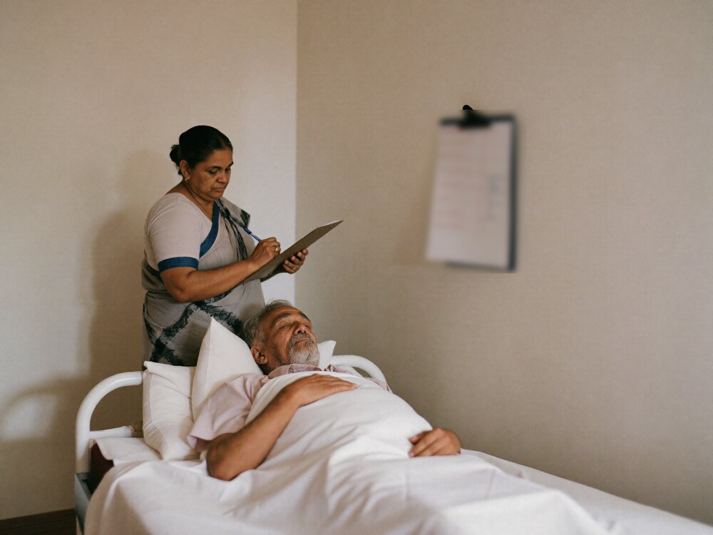 Family caregiver recording a turning time on a chart beside an elderly patient resting in bed
