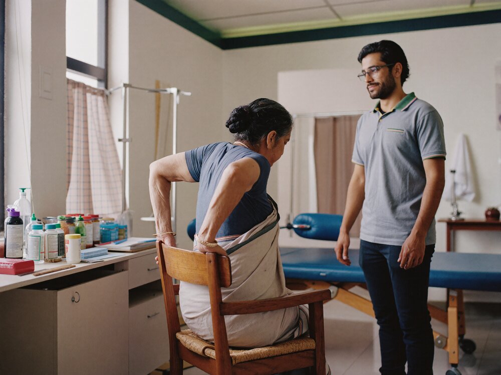 Older woman rising from a wooden chair in the sit-to-stand exercise while a physiotherapist observes