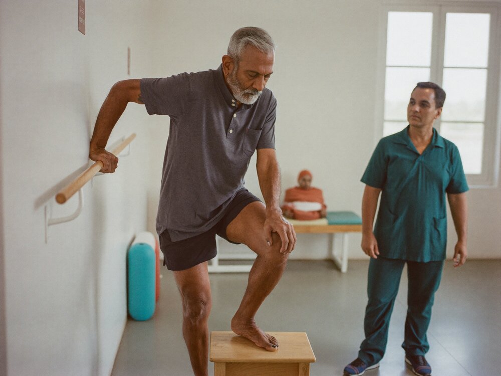 Older man stepping up onto a low wooden step with one hand on a rail while a physiotherapist watches