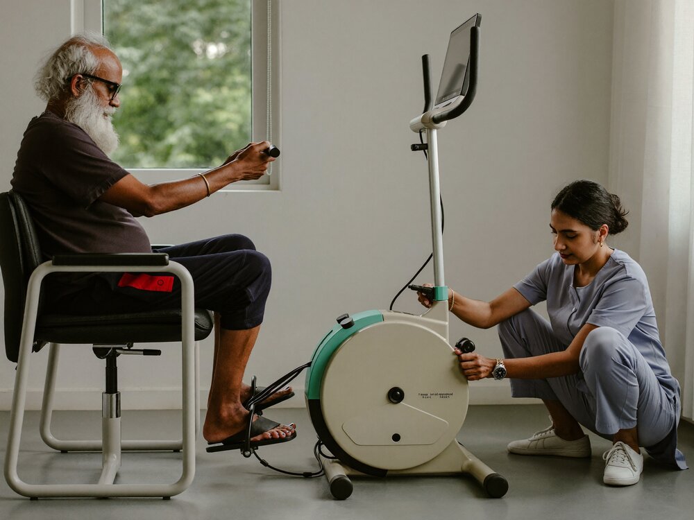 Elderly man pedalling a motorised leg cycle from a chair while a physiotherapist adjusts the settings