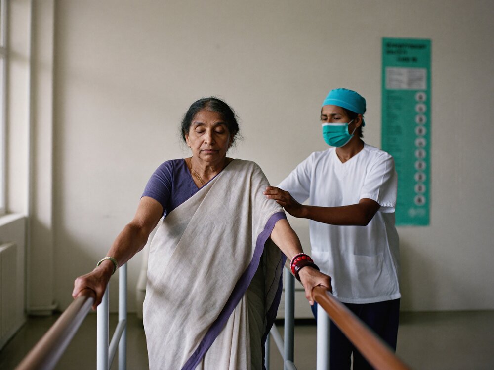 Older woman in a saree relearning to walk between parallel bars while a therapist guards her from behind